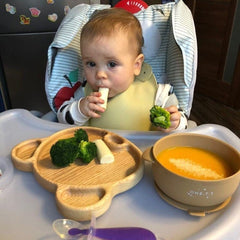 Baby in high chair eating from bear shaped wooden platter with broccoli