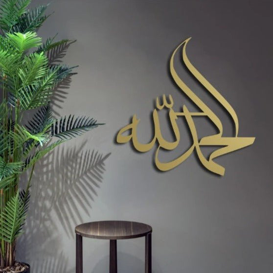 Gold Alhamdulillah Islamic calligraphy wall hanging on dark grey wall beside plant