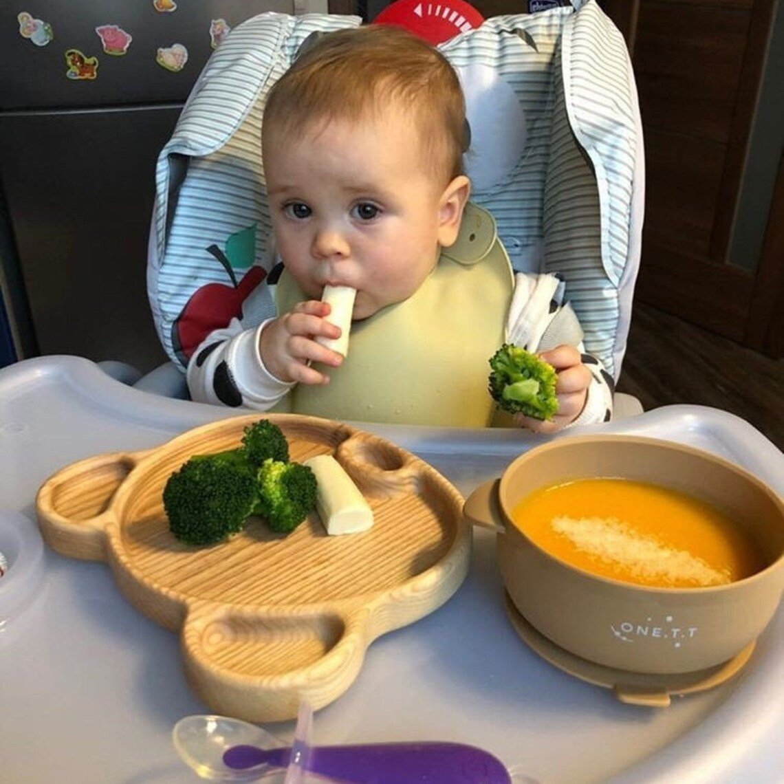 Baby in high chair eating from bear shaped wooden platter with broccoli