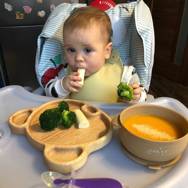 Baby in high chair eating from bear shaped wooden platter with broccoli