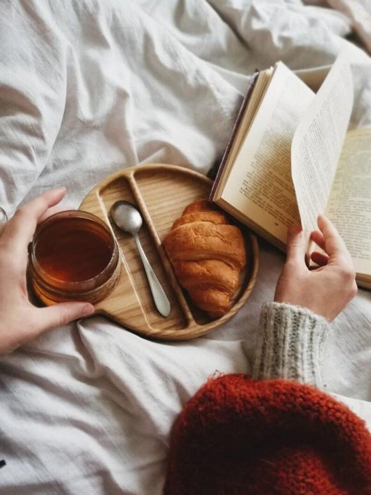 Person turning book page beside wooden food tray with tea and croissant