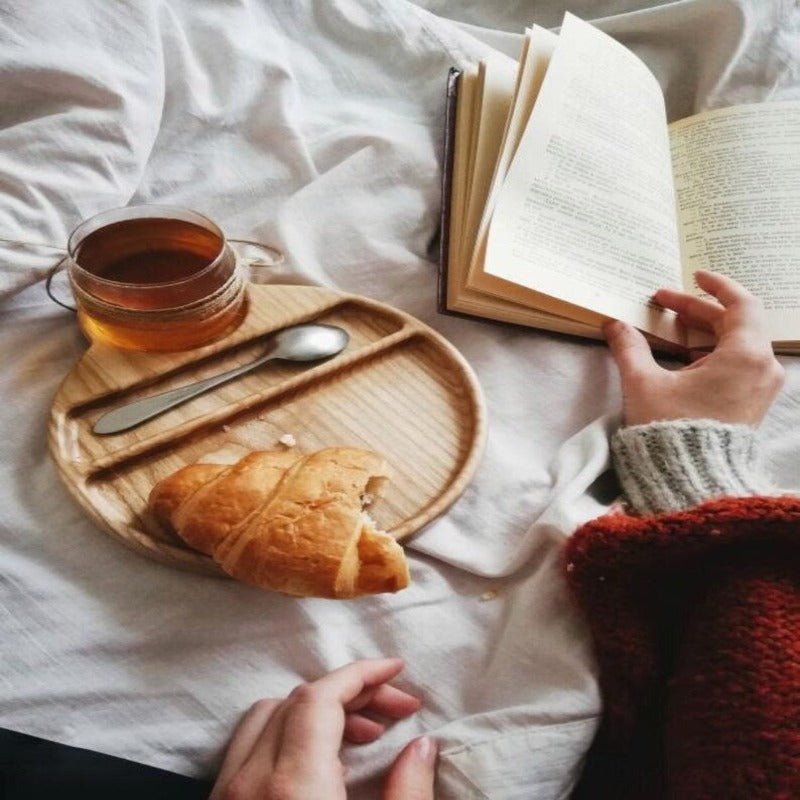 Wooden food tray with croissant, tea cup and spoon beside person reading