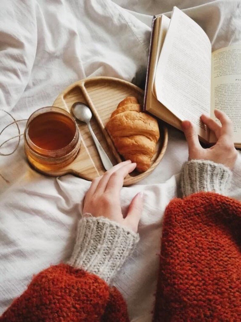 Hands holding wooden cup and spoon tray with croissant and tea on bed