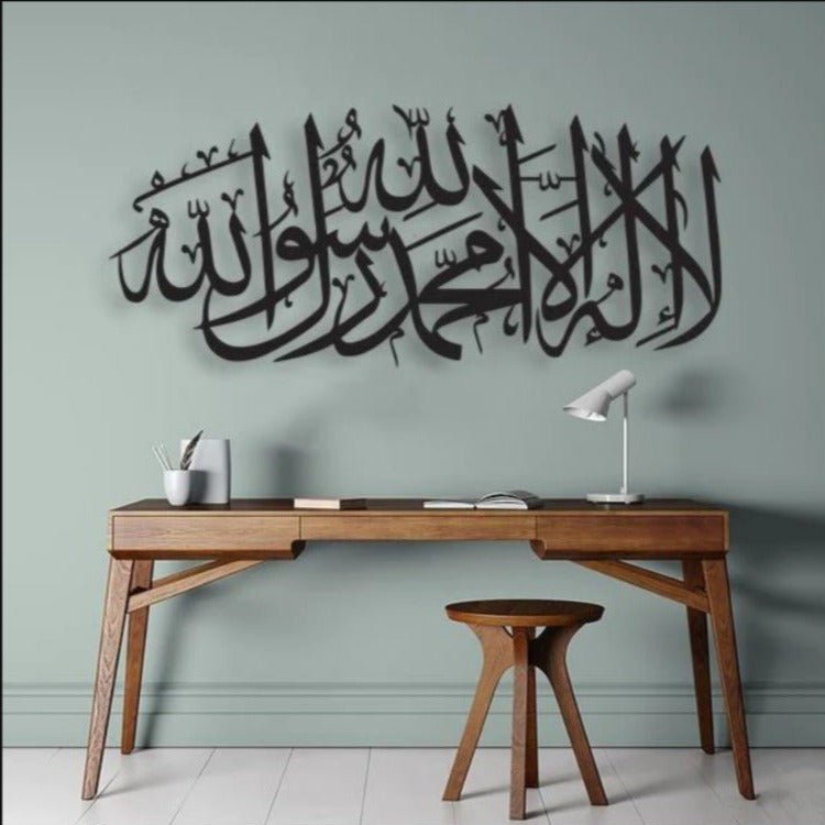Black horizontal Kalma Shareef calligraphy above wooden desk on grey green wall