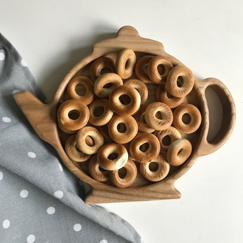 Teapot shaped wooden serving tray filled with ring biscuits on dotted cloth